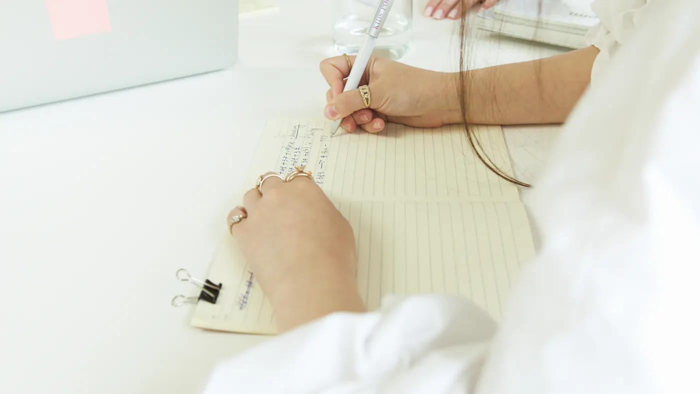 A hand writing in a notebook beside a laptop.